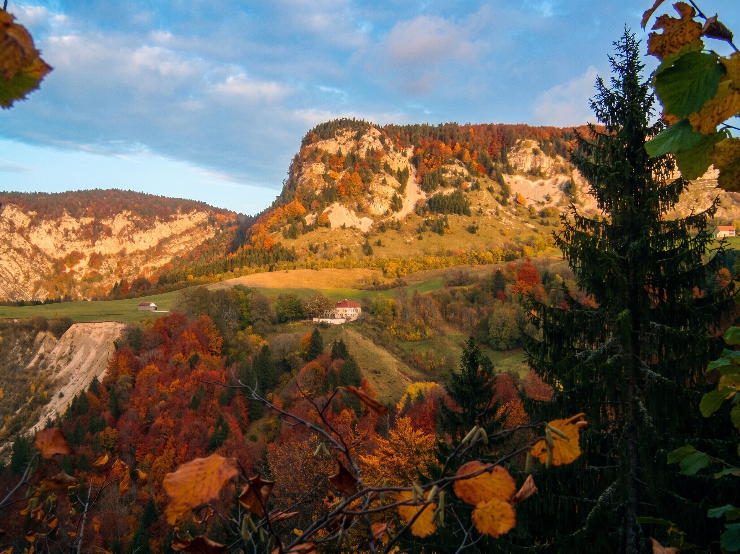 Paesaggio montano con fogliame autunnale, un piccolo edificio e cielo sereno.