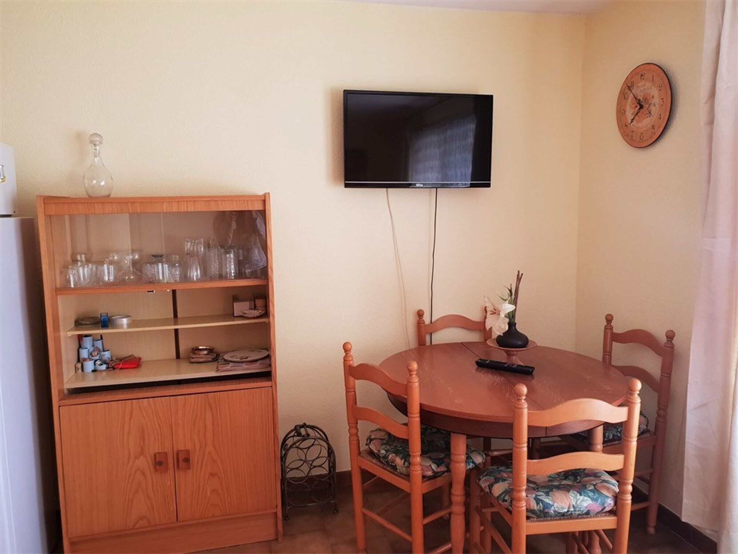 A dining area with a round wooden table, six chairs, a television mounted on the wall, a wooden cabinet with glass doors, and a clock on the wall.