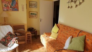 Een woonkamer met een rieten stoel, een bank met gekleurde kussens, een bijzettafeltje met een schaal, en een boekenplank.