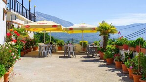 Un área de estar al aire libre con mesas y sillas bajo sombrillas amarillas, rodeada de plantas y flores en macetas, y una vista de montaña al fondo.