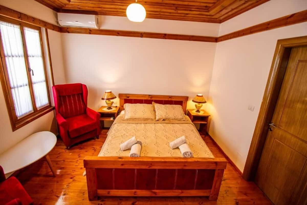 A wooden-framed bedroom with a bed, a red chair, bedside lamps, and a wooden door.