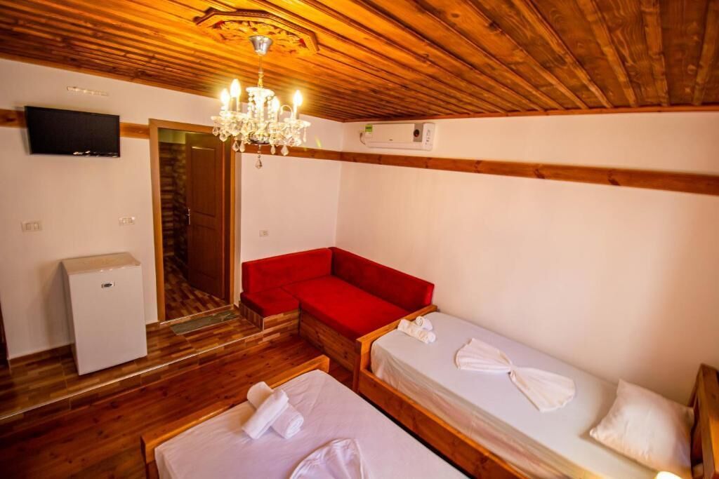 A room with wooden floors, a red sofa, two single beds with towels, a TV, a small white appliance, and a chandelier.