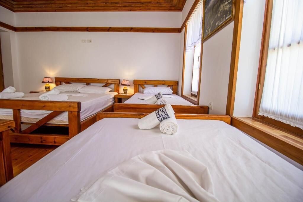 A hotel room with three wooden beds, each with a wooden headboard and white linens. There are two bedside tables with lamps, and a large window with curtains.