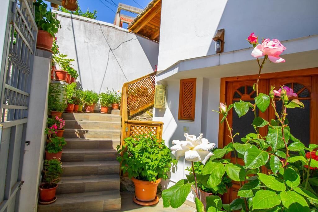 A narrow street with steps leading to a white building with a wooden door and window shutters. There are potted plants and flowers along the path.