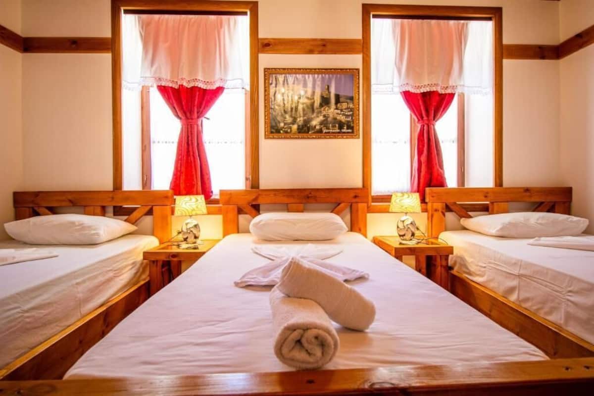 A hotel room with three wooden beds, each with white linens and pillows. There are two bedside tables with lamps and a framed picture on the wall between the windows.