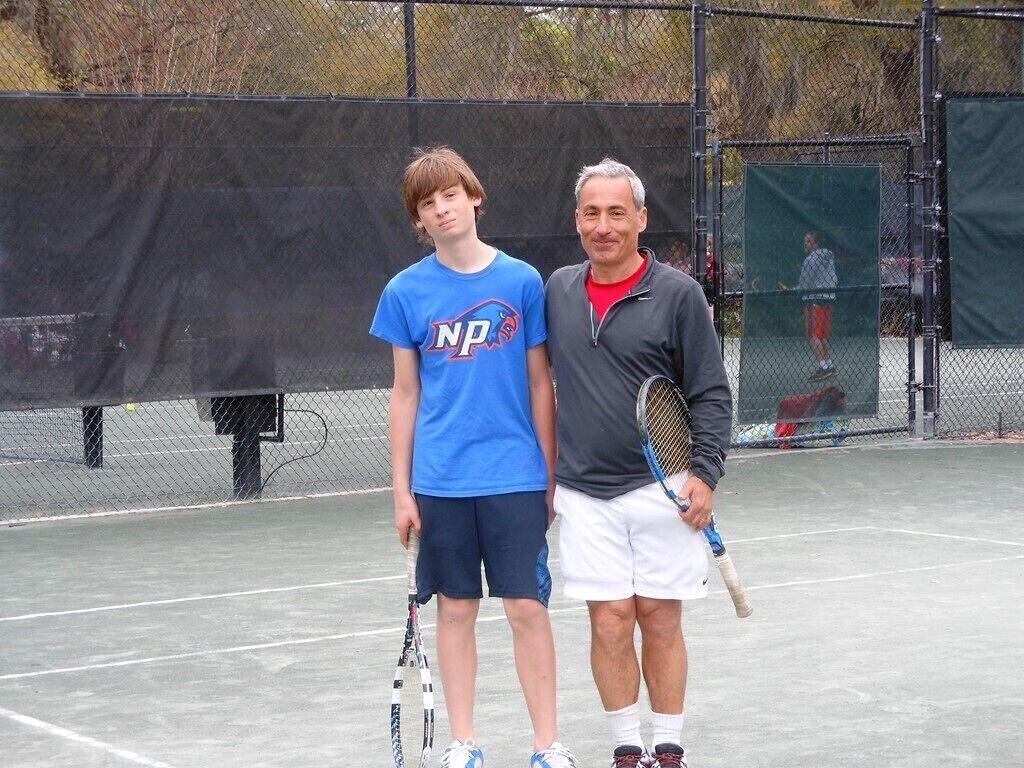 A tennis court with a chain-link fence, a net, and a tennis ball machine in the background.