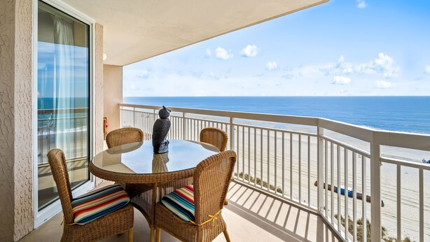 Ein Balkon mit Korbstühlen und einem Glastisch, mit Blick auf einen Strand.