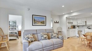 A modern living room with a grey sofa, wooden chairs, a dining table, and a kitchen area with white cabinets and appliances.
