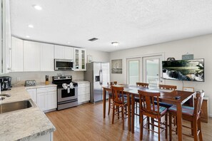 Una cucina con mobili bianchi, un frigorifero in acciaio inossidabile, un forno a microonde e un piano cottura. C'è un'area pranzo con tavolo e sedie in legno.