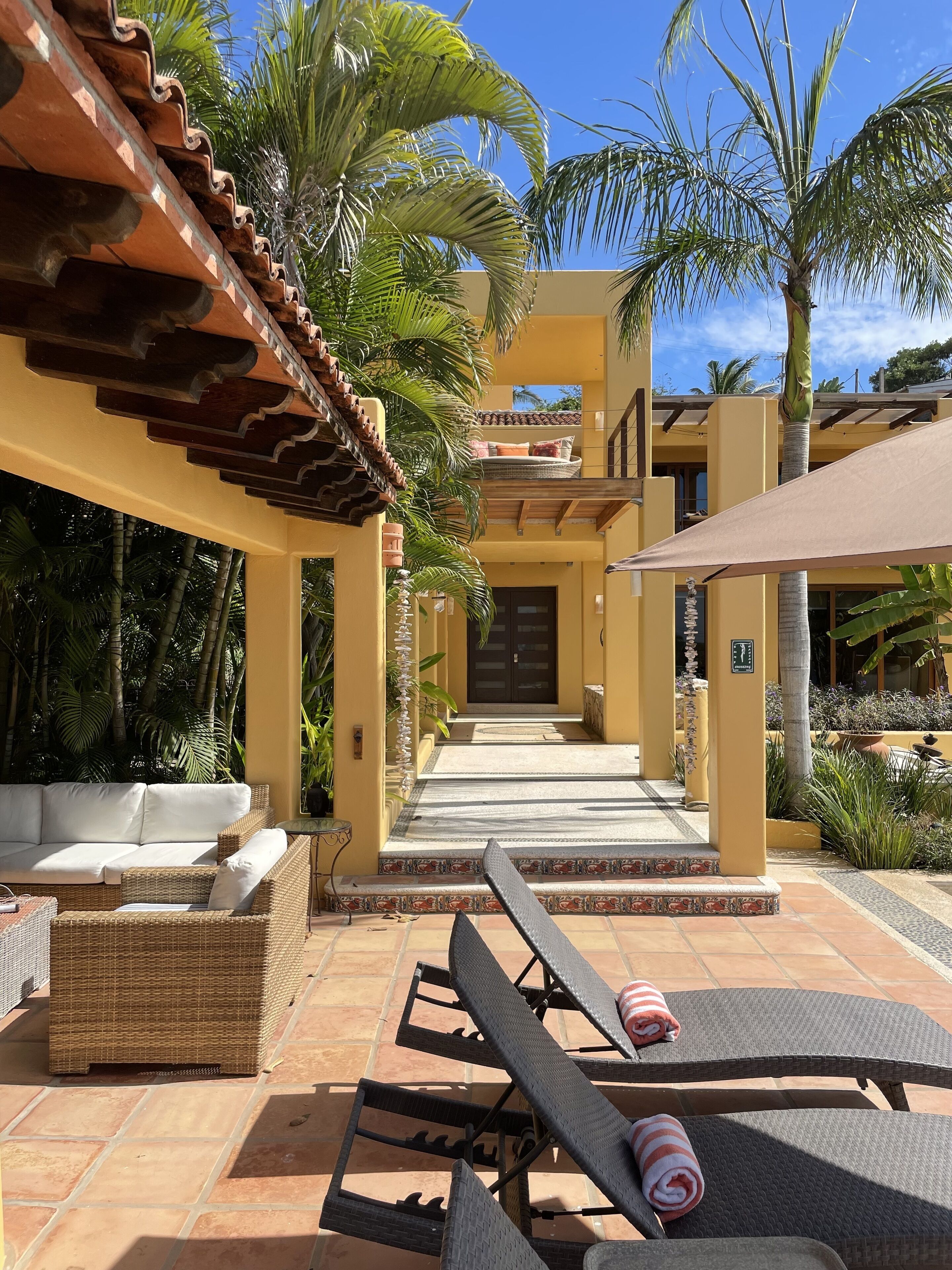 Un complejo turístico con un patio con mobiliario de mimbre y sillones de descanso, un camino de baldosas que conduce a la entrada y palmeras al fondo.