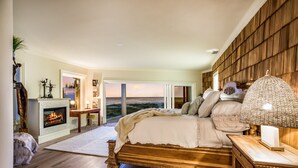 A bedroom with a large bed, a fireplace, a view of the ocean, and a wooden wall.