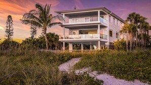 A two-story beachfront house with a balcony, surrounded by tropical vegetation and a sandy path leading to the entrance.
