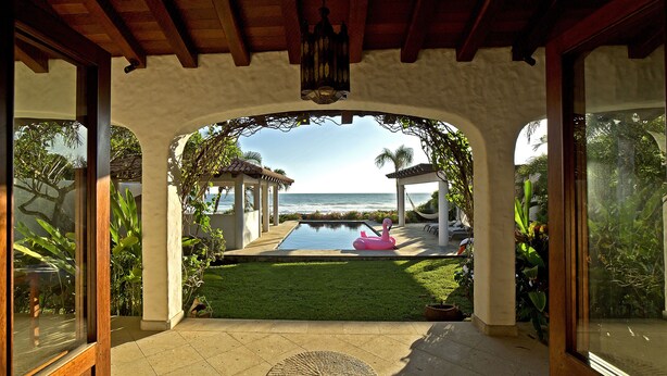 A covered patio with a view of the ocean, a pool, and a flamingo float.