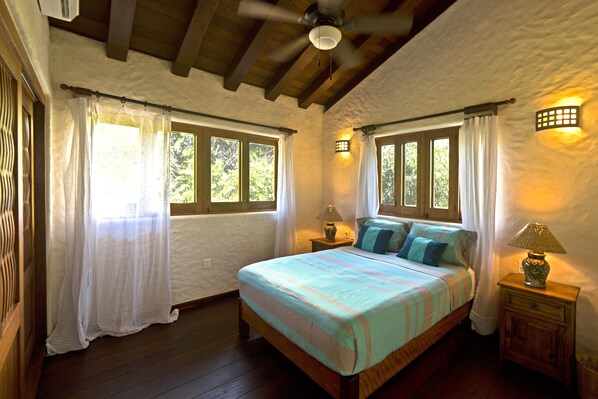 A bedroom with a wooden bed, two bedside tables, a ceiling fan, and windows with curtains.
