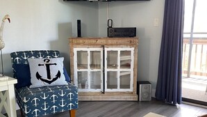 Una sala de estar con una televisión de pantalla plana montada en la pared, un mueble de madera con puertas de cristal, un sillón azul con estampado de anclas y una mesita con un objeto decorativo.