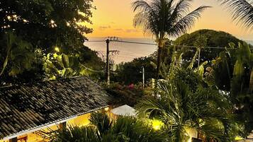 Uma área de piscina com vista para o mar ao pôr do sol, rodeada por vegetação tropical e um telhado tradicional em azulejos.