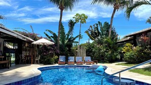 A swimming pool with lounge chairs and umbrellas, surrounded by tropical plants and palm trees.