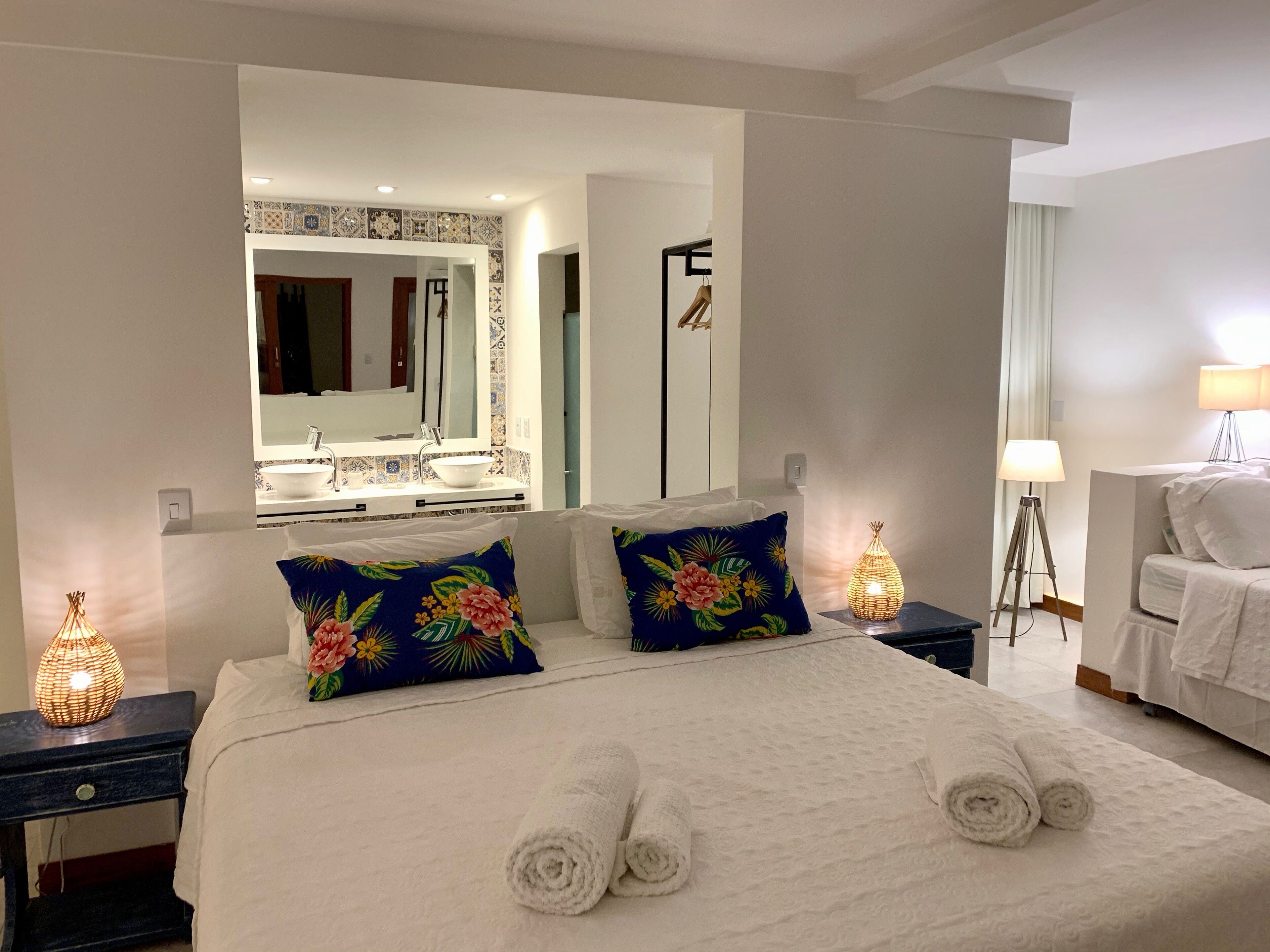 A hotel room with a large bed, two bedside tables, a nightstand, a mirror, a bathroom with a sink and mirror, and a lamp.