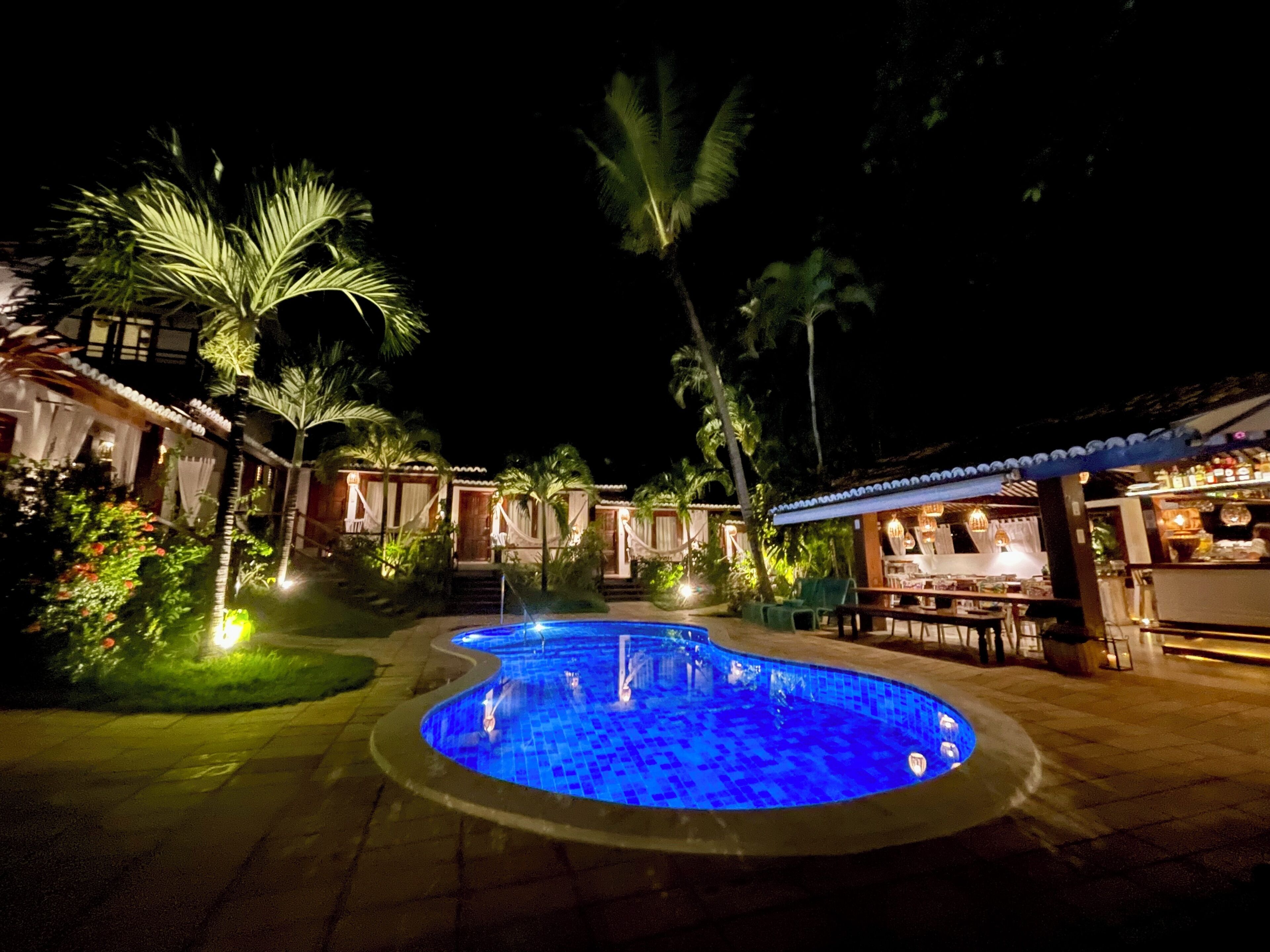 A nighttime pool area with a blue-lit pool, palm trees, and a bar area with outdoor seating.