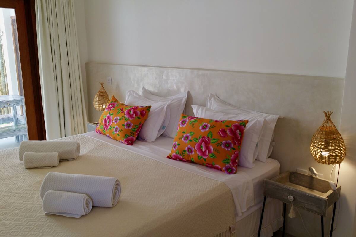 A neatly made bed with floral pillows, a bedside table with a lamp, and rolled towels.