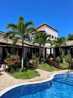 Zona junto a la piscina con piscina enlosada, palmeras y un edificio de techumbre y paredes blancas.