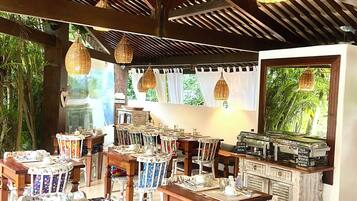 Un restaurant avec des tables et des chaises en bois, des lampes en osier suspendues et une vue sur la verdure environnante.