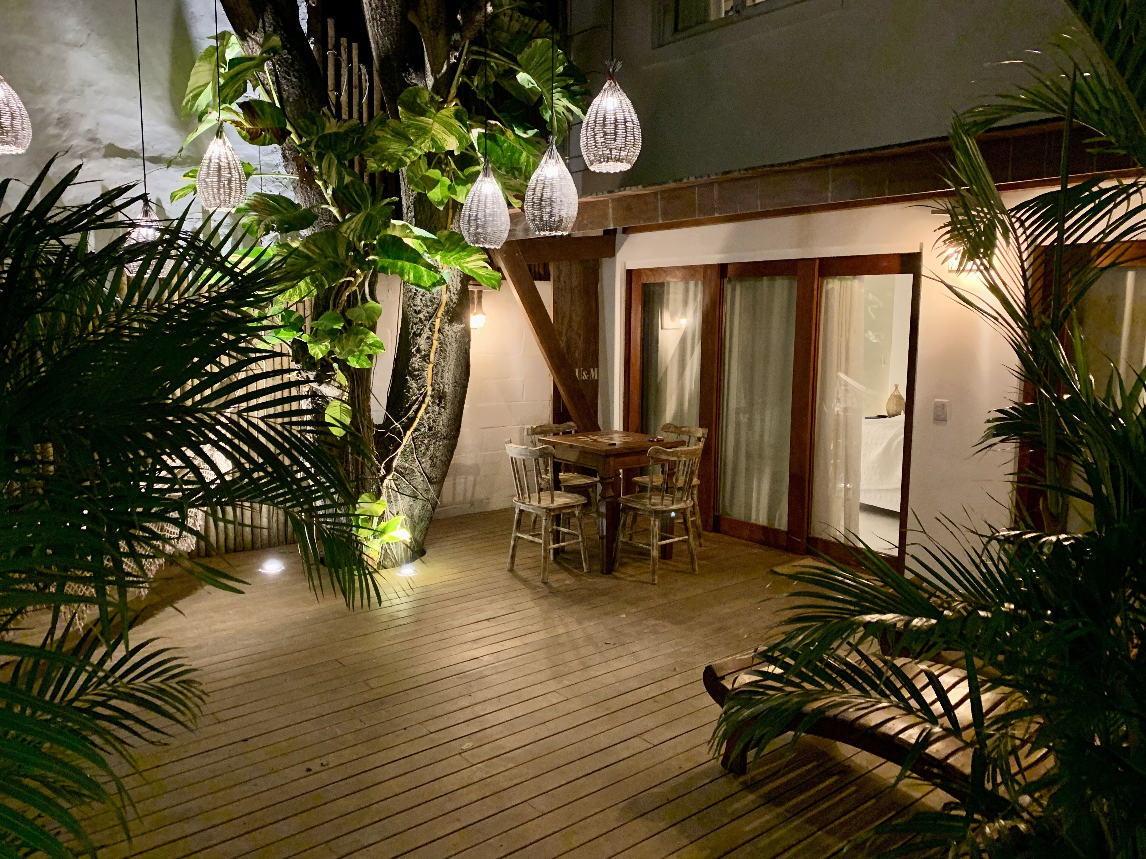 An outdoor patio area with wooden flooring, a table with chairs, hanging pendant lights, and lush green plants.