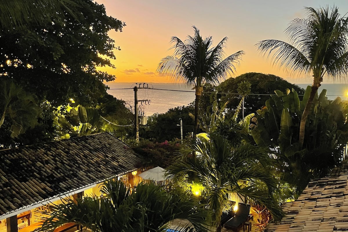 A tropical landscape with a view of the sea at sunset, palm trees, and traditional tiled roofs.