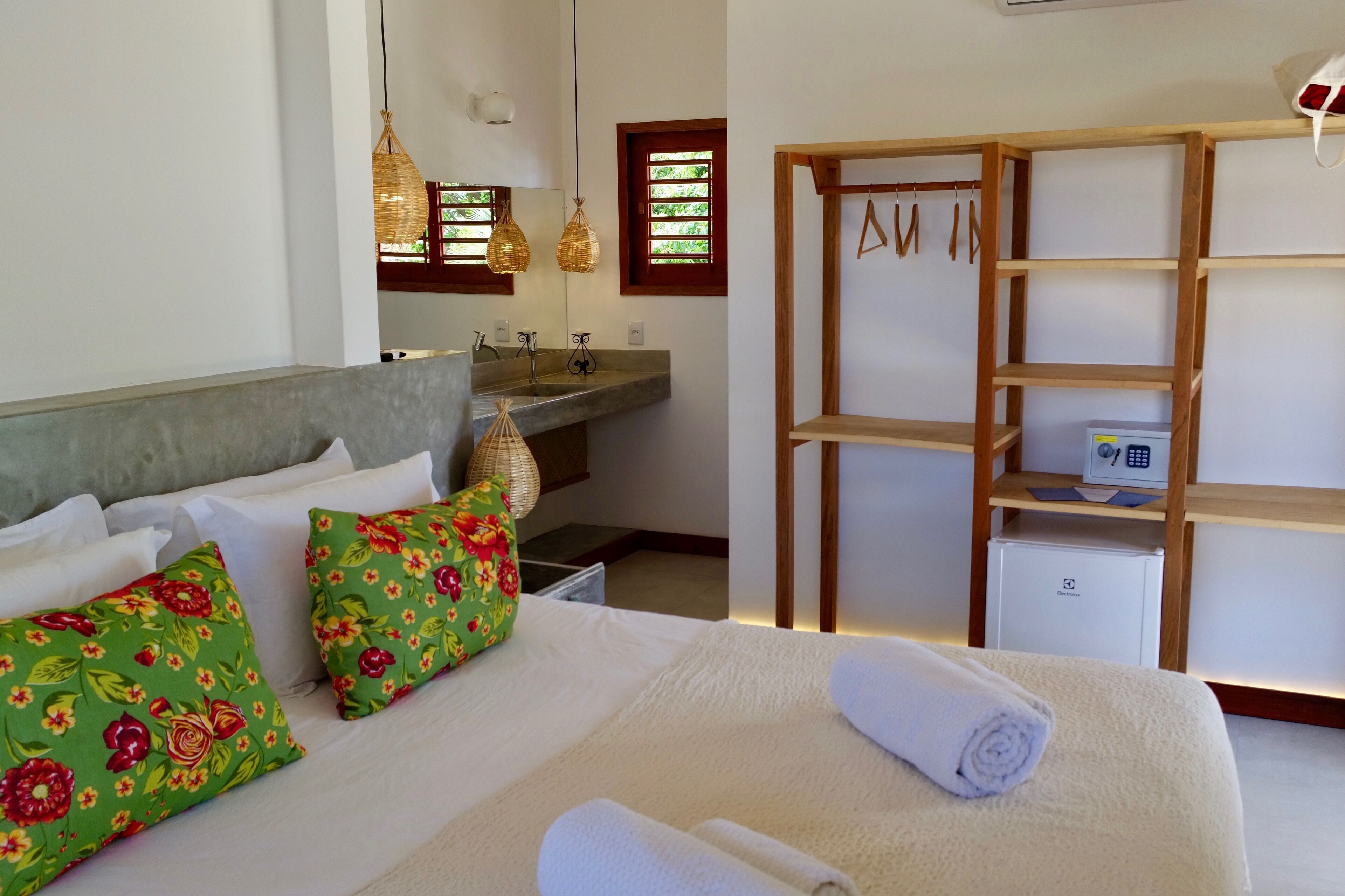 Una habitación de hotel con una cama, almohadas, un estante de madera, un escritorio con computadora y una ventana con persianas.