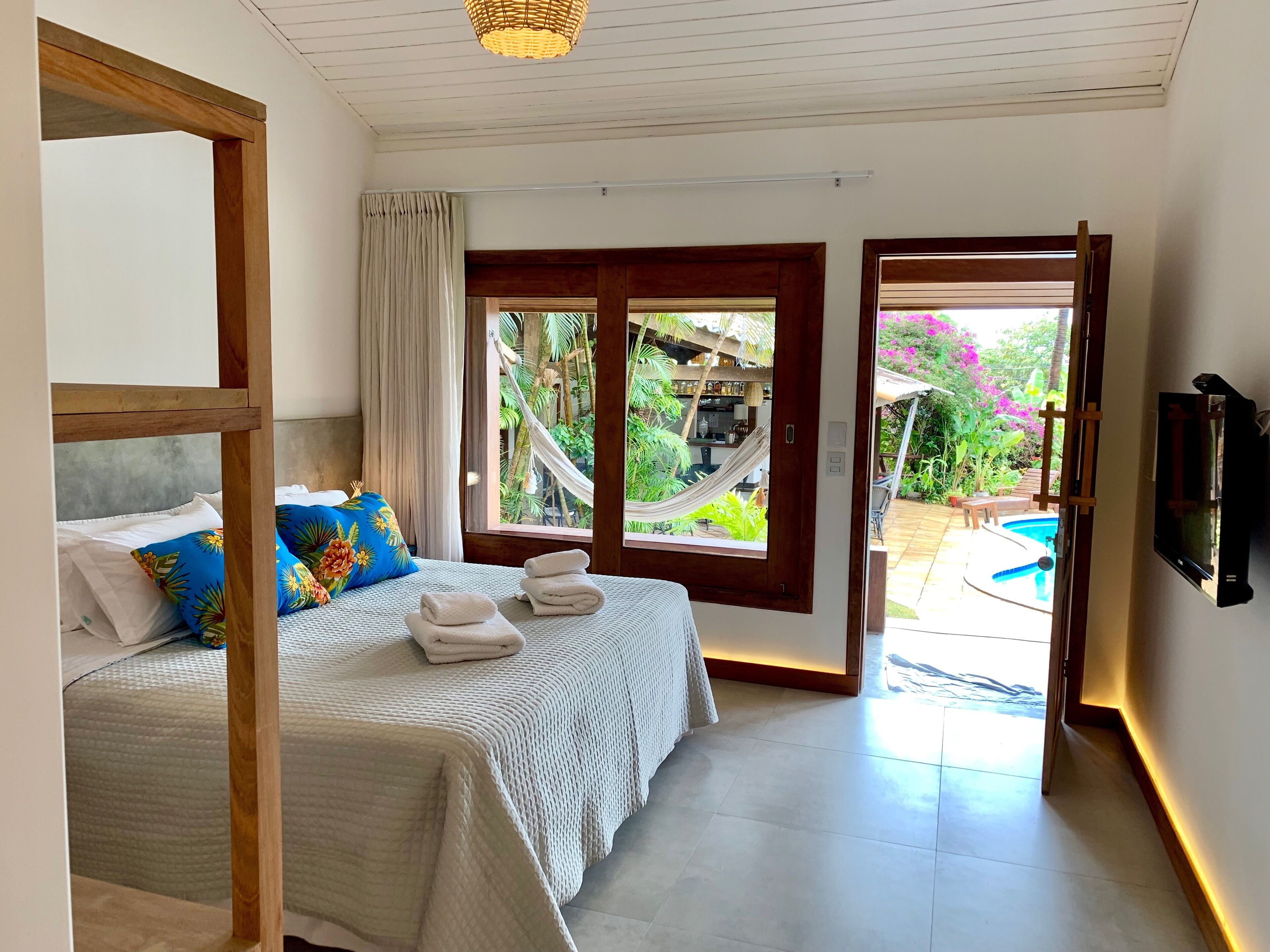 Un dormitorio con cama, almohadas, televisor y vistas a una piscina y jardín.