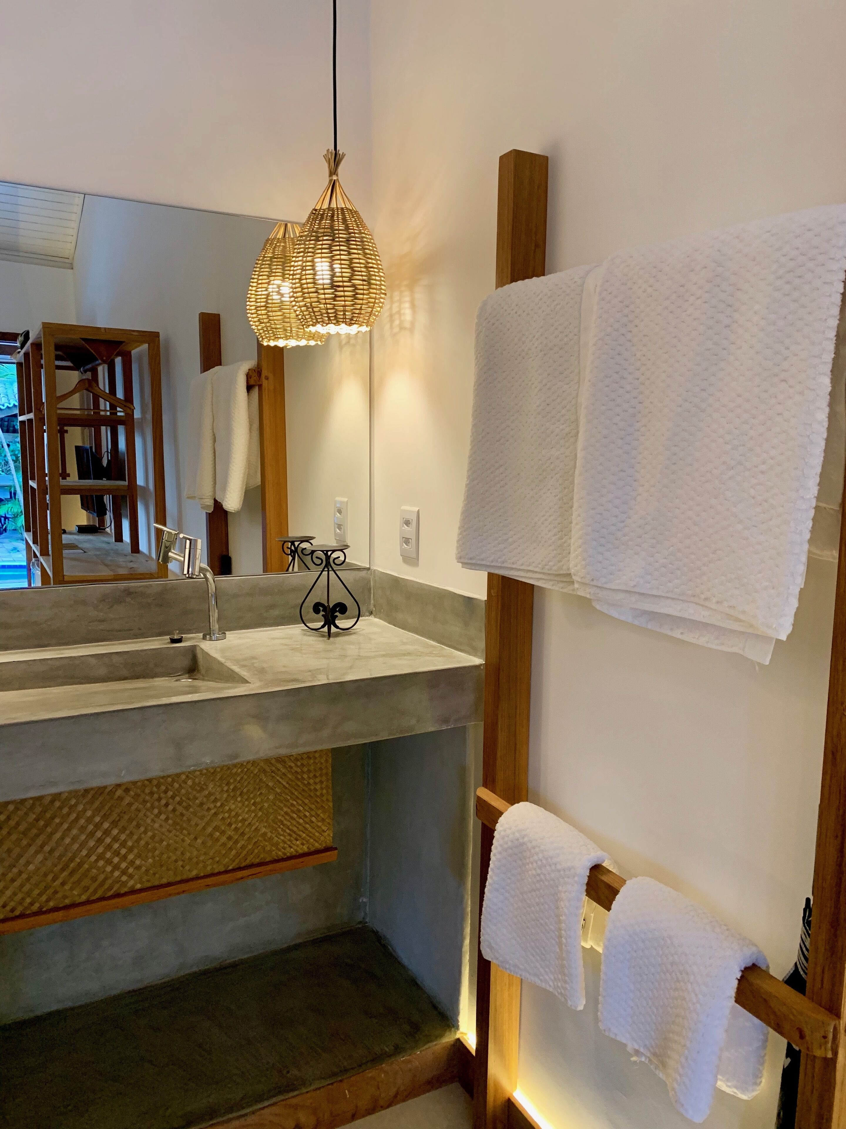 A bathroom with a large mirror, a wooden towel rack, a concrete sink, and a woven light fixture.