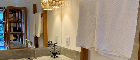A bathroom with a large mirror, a wooden towel rack, a concrete sink, and a woven light fixture.