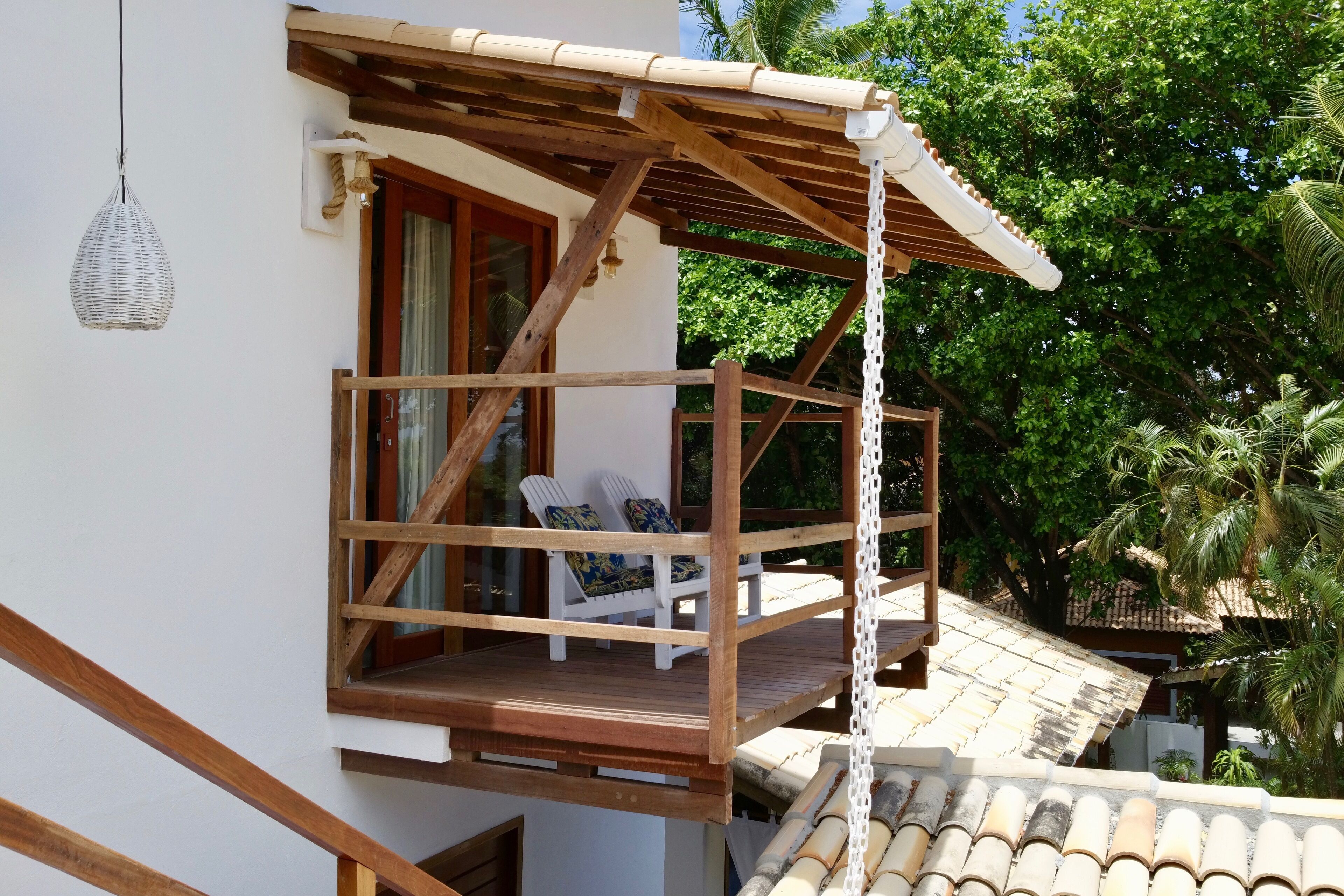 A wooden balcony with two Adirondack chairs, a hanging light fixture, and a view of palm trees.