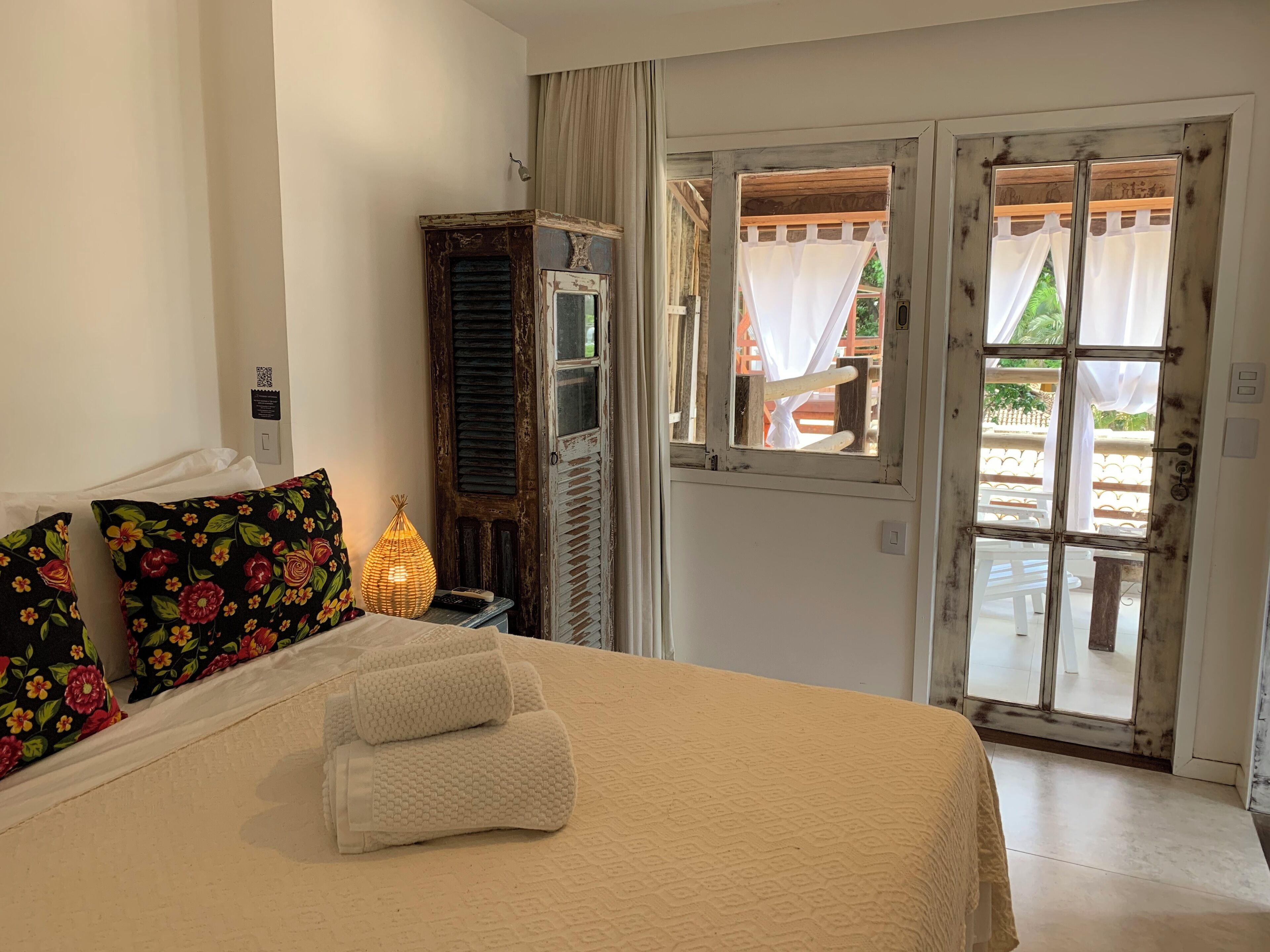 A bedroom with a bed, a nightstand, a lamp, a wooden wardrobe, and a view of the outside through a glass door.