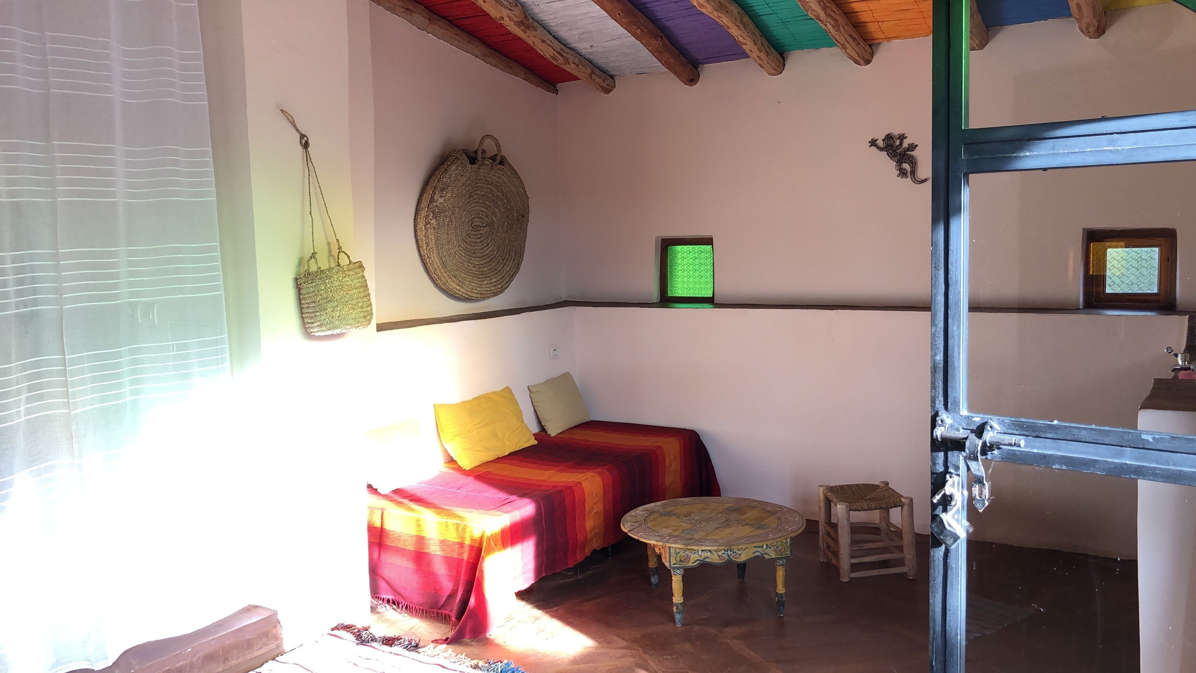 A cozy room with a bed, a small table, a woven bag, and a framed picture on the wall.