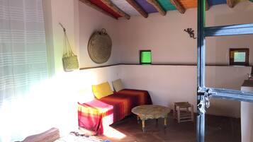 A cozy room with a bed, a small table, a woven bag, and a framed picture on the wall.