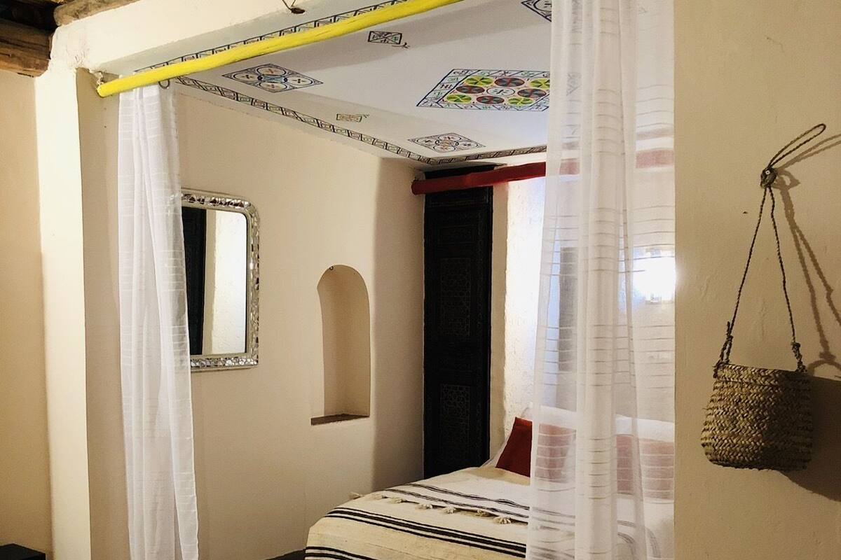 Ein Zimmer mit einem Bett, das unter einem Baldachin steht, einem Spiegel, einem kleinen Tisch und einem hängenden Korb.