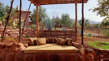 Un área de estar al aire libre con techo de paja, rodeada de plantas desérticas y un muro de piedra.