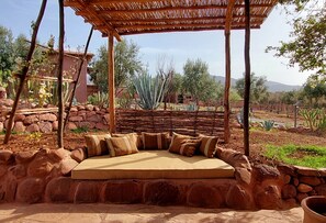 Un espace extérieur aménagé avec des sièges, doté d’un toit de chaume et entouré de plantes désertiques et d’un mur en pierre.