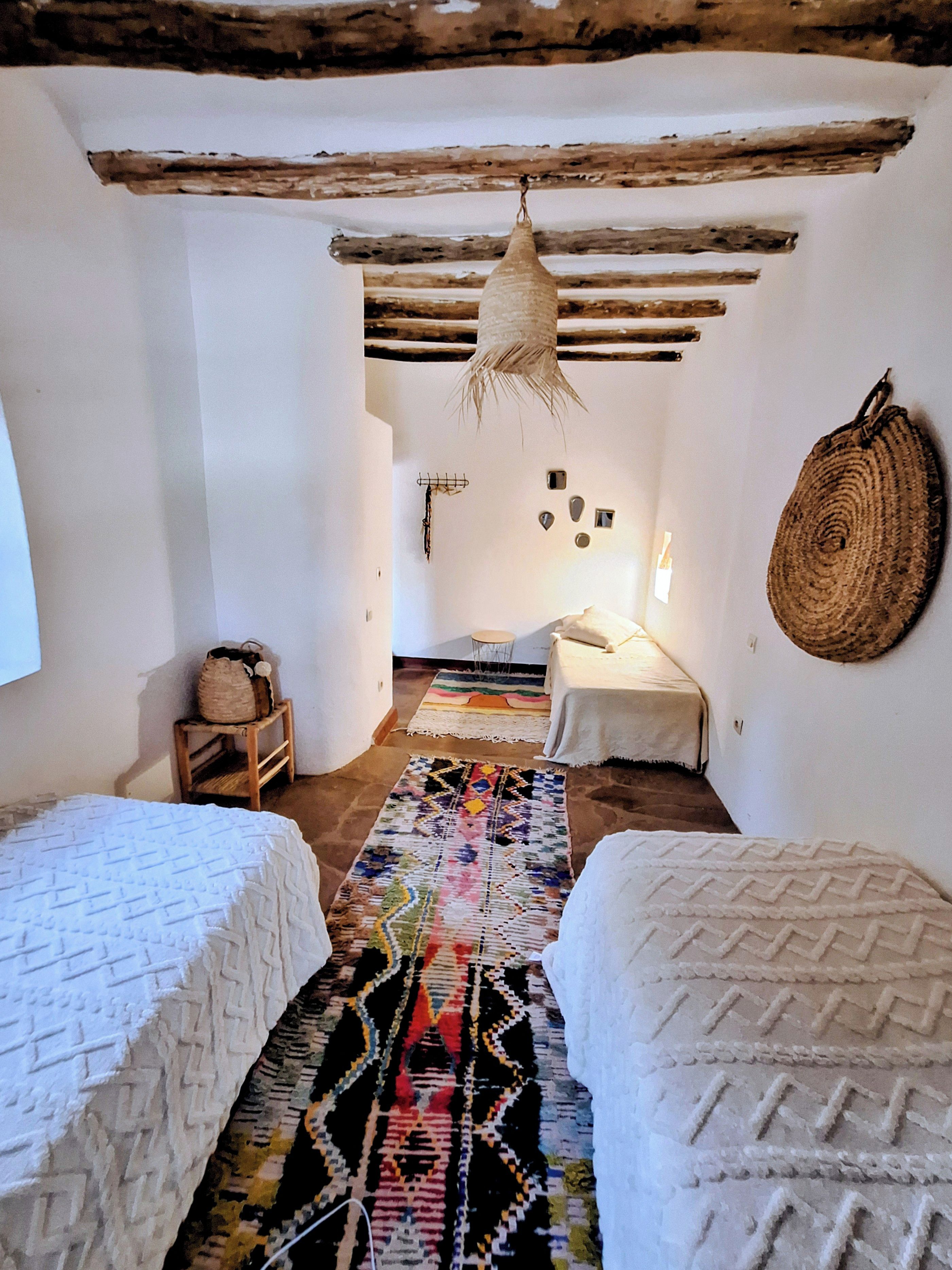 A room with two beds, a hanging light fixture, a woven wall hanging, and a colorful rug.