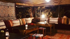 Une tente chaleureuse et faiblement éclairée, ornée d’une tenture murale à motifs, avec une table en bois et un coin salon confortable doté de coussins.