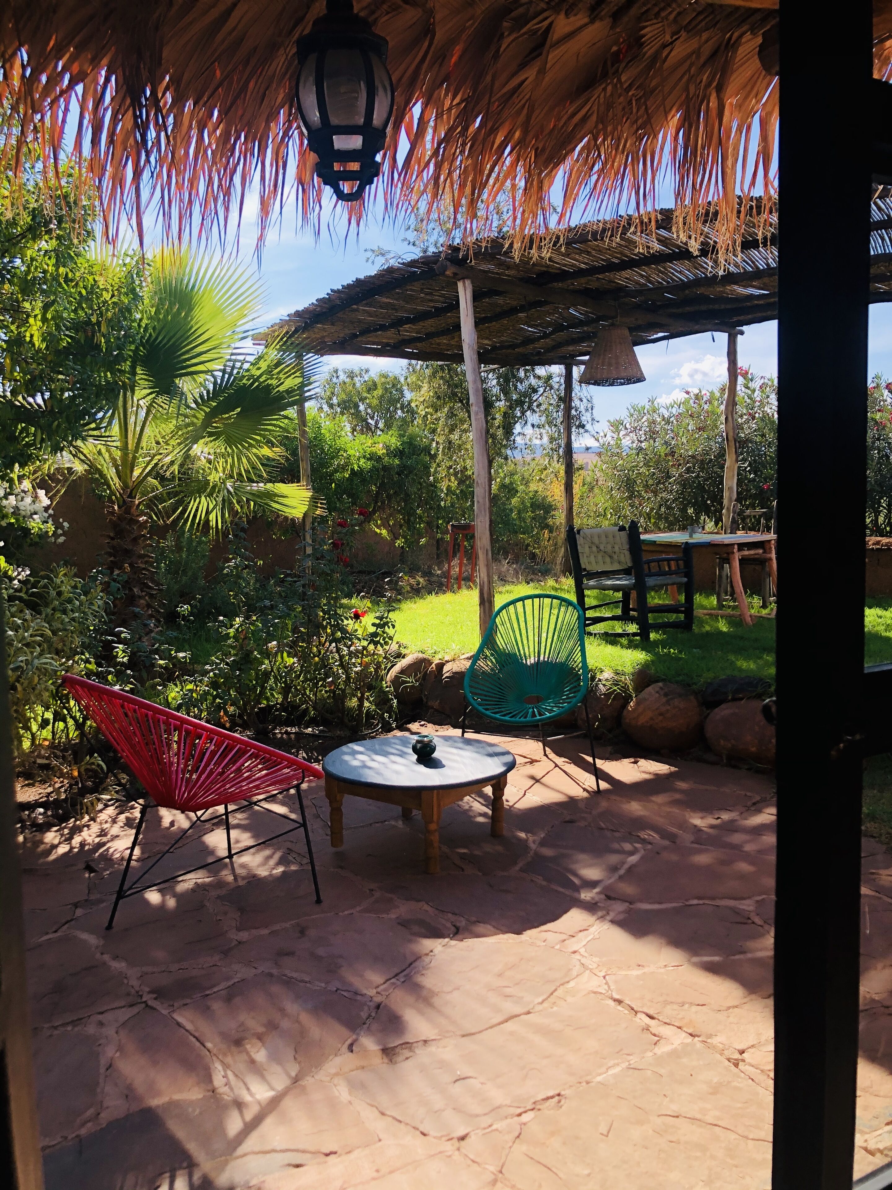 Un patio al aire libre con techo de paja, dos sillas, una mesa y un farol colgante.