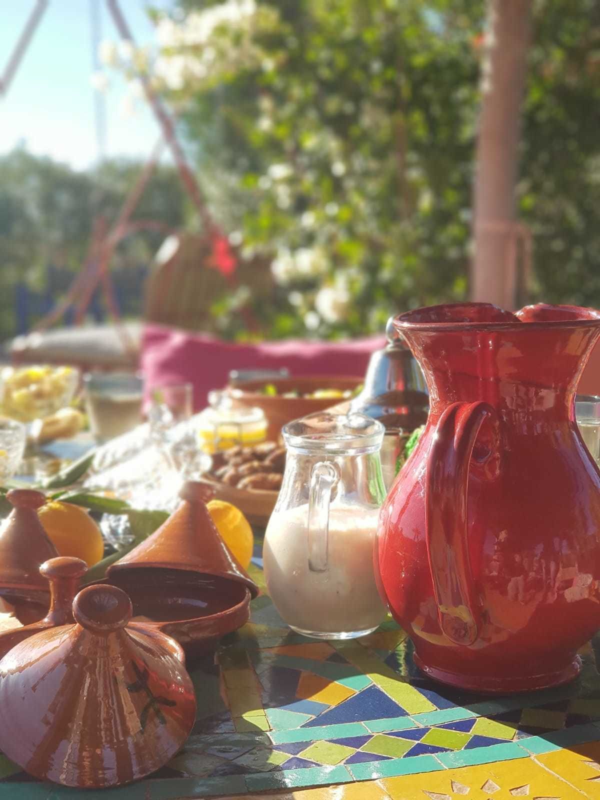 A table with various kitchenware, including a red pitcher, a glass jug, and terracotta pots, set outdoors with a colorful tiled table and greenery in the background.