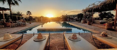 A pool area with lounge chairs and tables, a clear view of the sunset, and a calm water surface reflecting the sky.