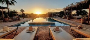 Un espace piscine doté de chaises longues et de tables, offrant une vue dégagée sur le coucher du soleil, et une surface d’eau calme qui reflète le ciel.