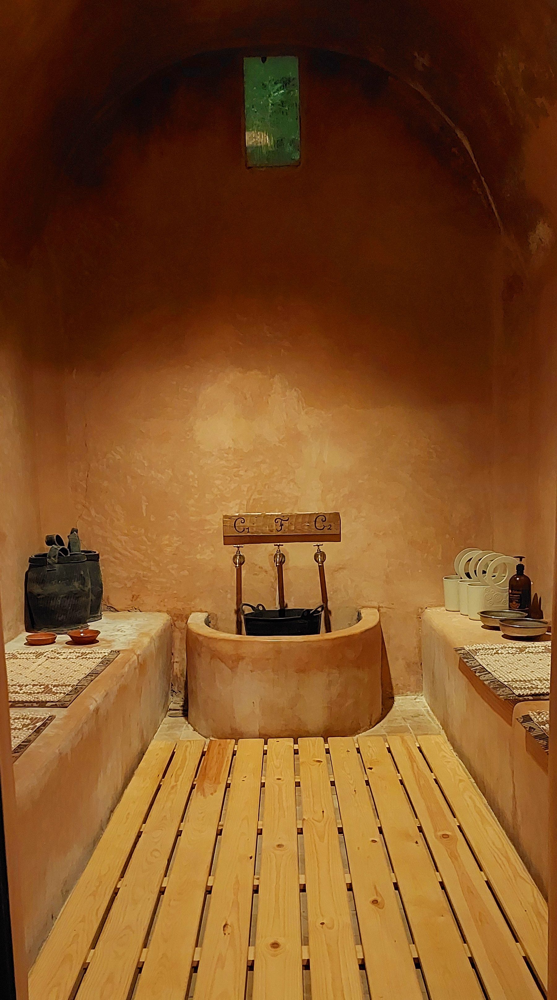 Una habitación con un banco de madera, un lavamanos pequeño y un estante con objetos.