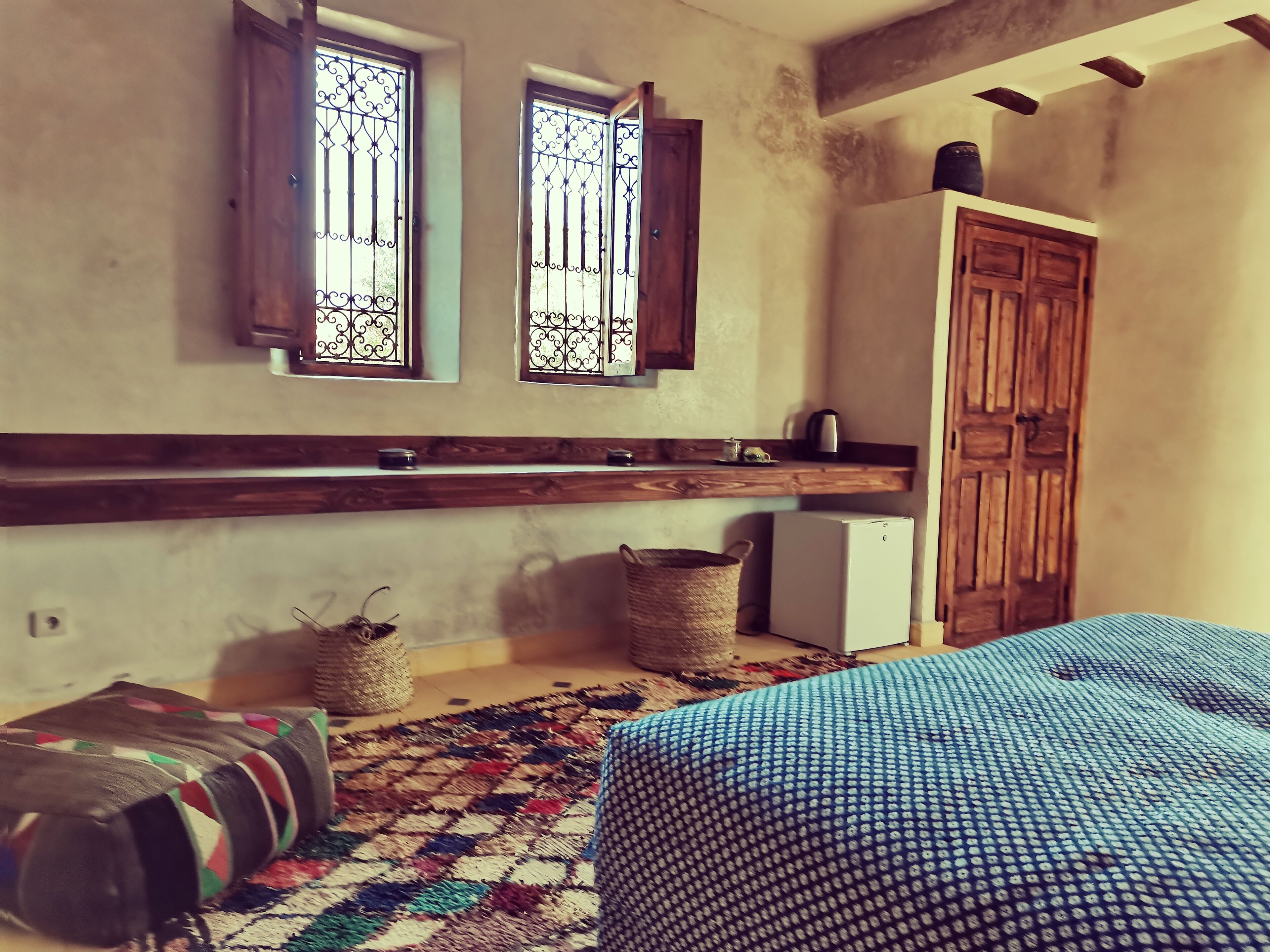 A room with a bed, a small kitchenette, a wooden wardrobe, and a window with bars.