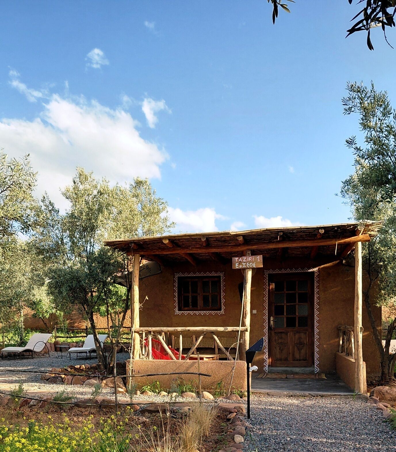 A rustic building with a porch, a wooden door, and a sign indicating 'TAZIRI 1'.