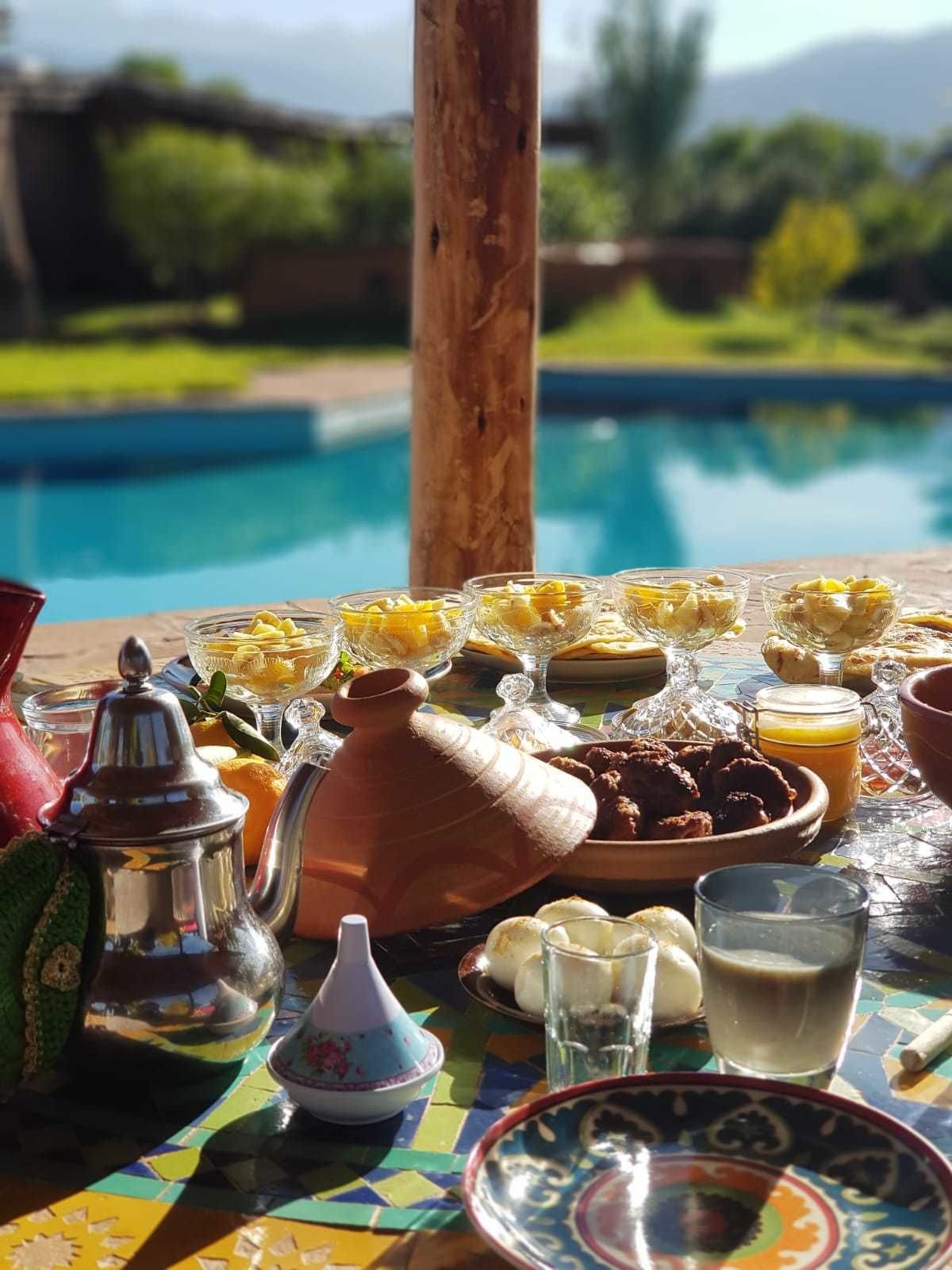 A table set with various dishes and drinks, a teapot, and a traditional Moroccan tagine, overlooking a pool.
