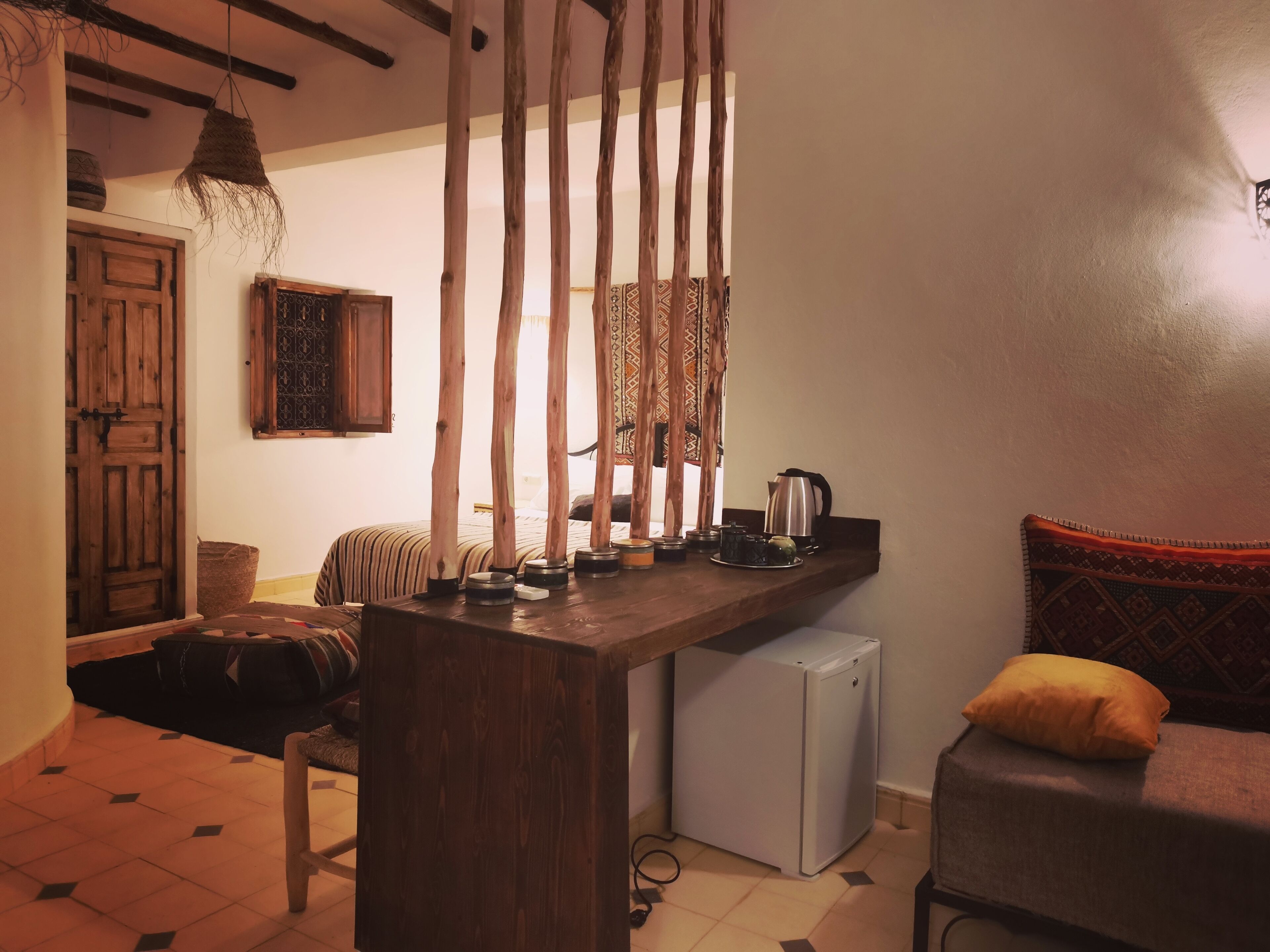 Una habitación acogedora con un escritorio de madera, un refrigerador pequeño, una cama con ropa de cama estampada y una lámpara colgante.
